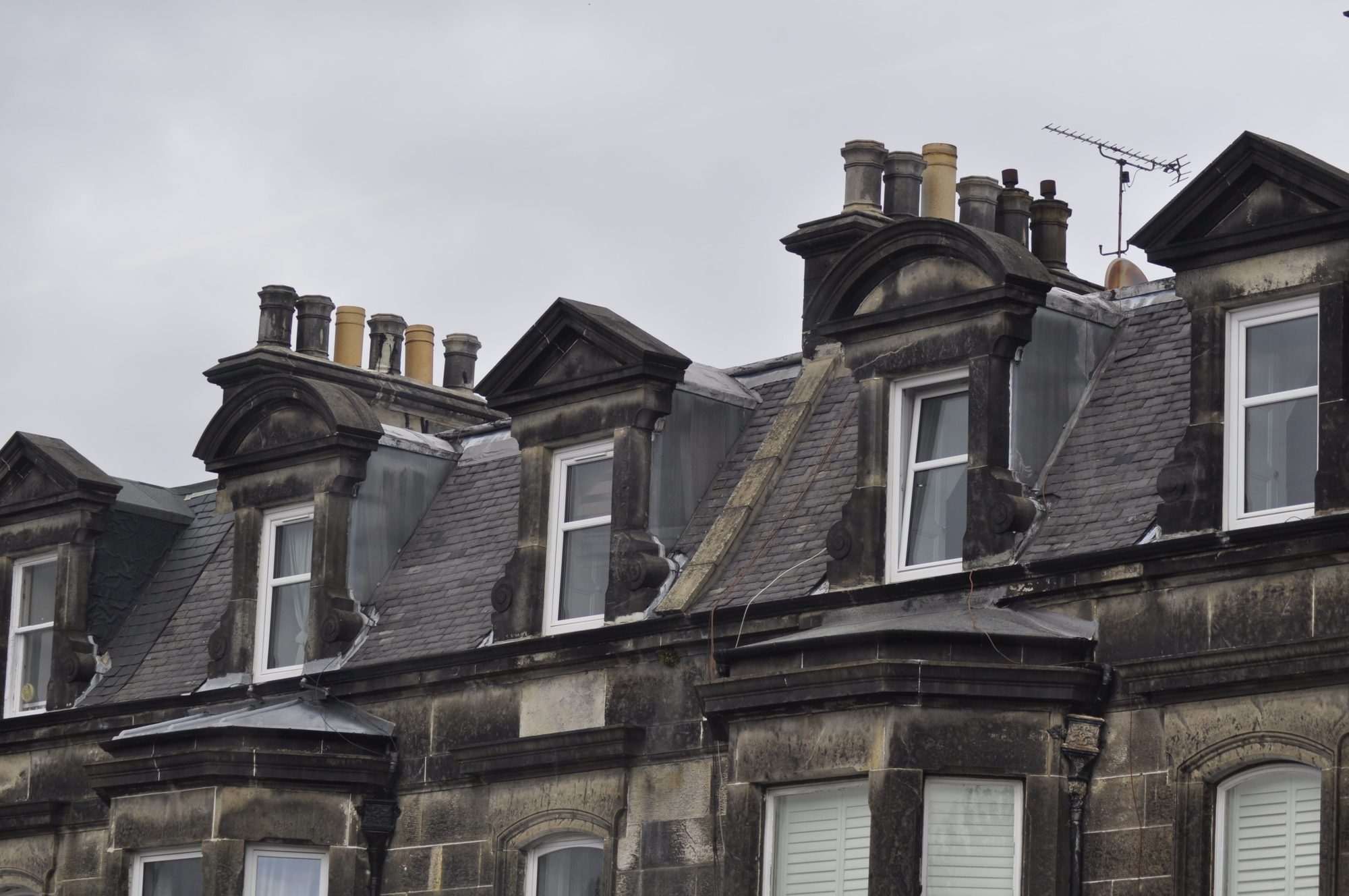 A close up row of chimney stacks and slate dormers, showing lead flashing and coursing detail around each junction
