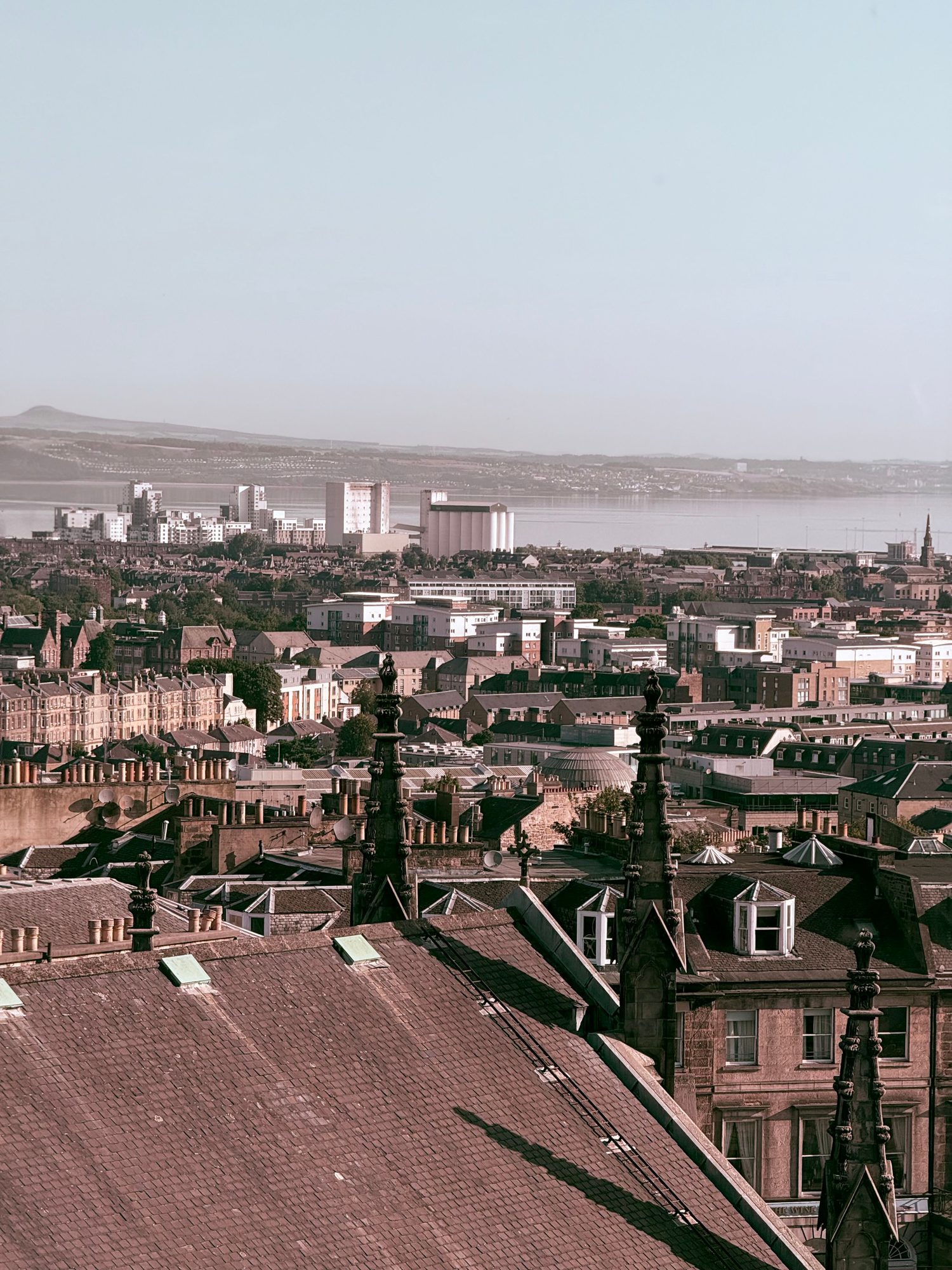 A rooftop view over ornate spires with the Firth visible in the distance, part of Edinburgh's historic skyline